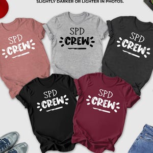May include: A collection of t-shirts in various colors, including heather mauve, athletic heather, dark grey heather, black, and maroon. Each shirt displays the text "SPD CREW" in a white, stylized font. A red baseball cap, sunglasses, and jeans are also visible.