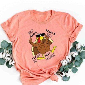 May include: A peach-colored t-shirt with a cartoon turkey wearing sunglasses and holding a plate of food. The text on the shirt reads "Turkey Gravy, Beans & Rolls, Let Me See That Casserole".