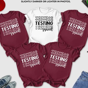 May include: A collection of maroon and white t-shirts with the text "Testing Squad" in a stacked design. A red baseball cap, blue jeans, sunglasses, and sneakers are also visible. The text "Shirt colors may look slightly darker or lighter in photos" is at the top.