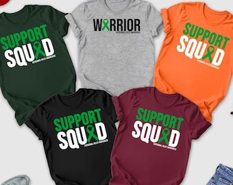 May include: A collection of t-shirts in various colors, including forest green, orange, black, and maroon. Each shirt features the words "SUPPORT SQUAD" in white with a green ribbon graphic. A gray shirt reads "WARRIOR". A red baseball cap and sunglasses are also visible.