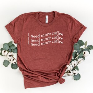 Puede incluir: Una camiseta marr&oacute;n con el texto "I need more coffee" repetido cuatro veces en blanco.