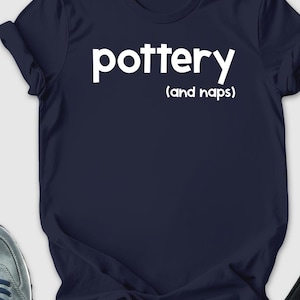May include: Navy blue t-shirt with the word "pottery" and "(and naps)" printed in white. The image also includes a straw hat, sunglasses, a wallet, sneakers, black pants, and a succulent plant in a white pot.