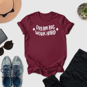 A maroon t-shirt with the text "DREAM BIG WORK HARD" printed on it, along with a pair of sunglasses, a hat, a wallet, and a potted succulent plant.