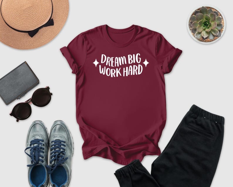 A maroon t-shirt with the text "DREAM BIG WORK HARD" printed on it, along with a pair of sunglasses, a hat, a wallet, and a potted succulent plant.