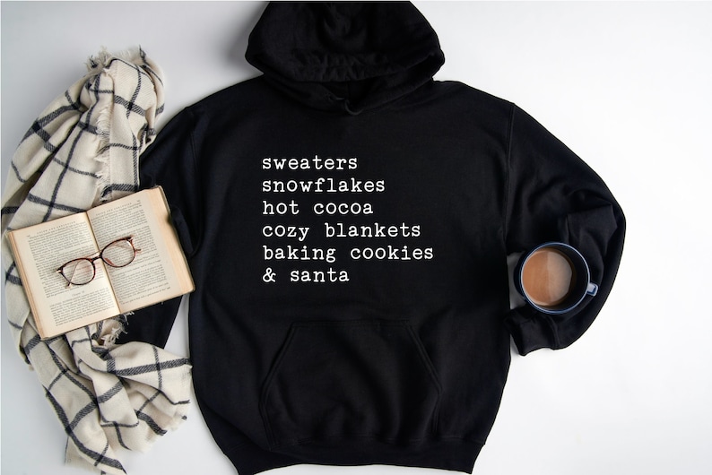 May include: Black hoodie with white text that reads "sweaters, snowflakes, hot cocoa, cozy blankets, baking cookies & santa".
