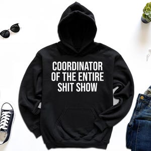 A black hoodie with white text, a pair of black and white sneakers, a pair of blue jeans, a watch, sunglasses, and a potted plant are arranged on a white background.
