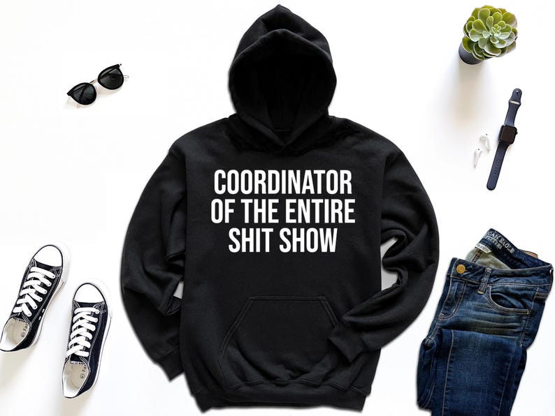 A black hoodie with white text, a pair of black and white sneakers, a pair of blue jeans, a watch, sunglasses, and a potted plant are arranged on a white background.