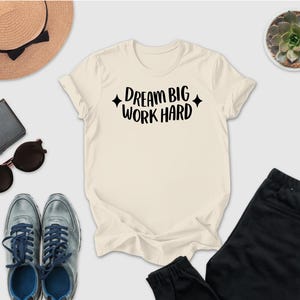 a casual outfit consisting of a white t-shirt with the text "DREAM BIG WORK HARD" printed on it, a pair of gray sneakers, a hat, sunglasses, a wallet, a potted succulent plant, and a pair of black pants.