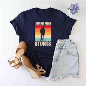 May include: A navy blue t-shirt with a retro-style graphic of a person on crutches. The graphic has a rainbow color scheme and the text "I do my own stunts".