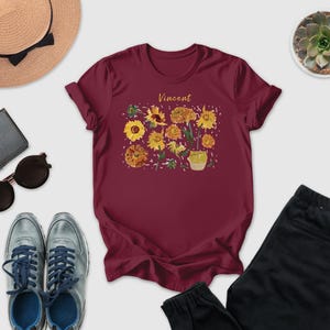 a casual outfit consisting of a burgundy t-shirt with a floral design, a pair of sunglasses, a hat, a wallet, a pair of sneakers, and a pair of pants.