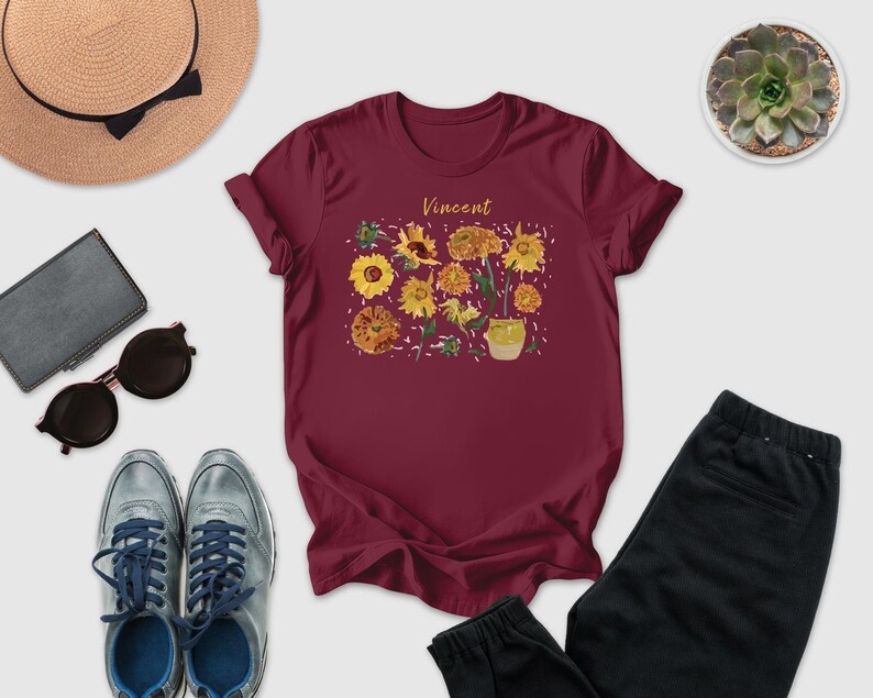 a casual outfit consisting of a burgundy t-shirt with a floral design, a pair of sunglasses, a hat, a wallet, a pair of sneakers, and a pair of pants.