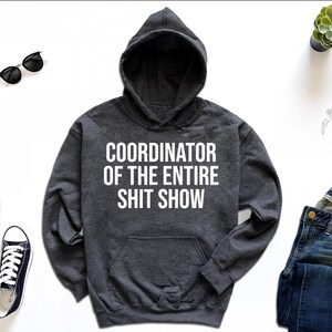 A gray hoodie with the text "COORDINATOR OF THE ENTIRE SHIT SHOW" printed on it, along with a pair of jeans, sneakers, a watch, and sunglasses.