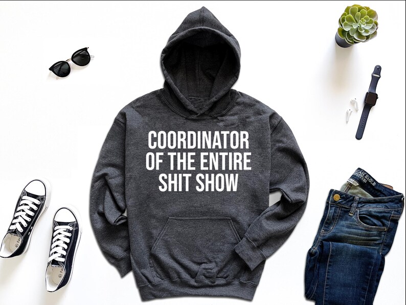 A gray hoodie with the text "COORDINATOR OF THE ENTIRE SHIT SHOW" printed on it, along with a pair of jeans, sneakers, a watch, and sunglasses.