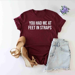 May include: Burgundy t-shirt with white text that reads "You had me at feet in straps". The shirt is laid flat on a white surface with a pair of brown sandals and denim shorts.