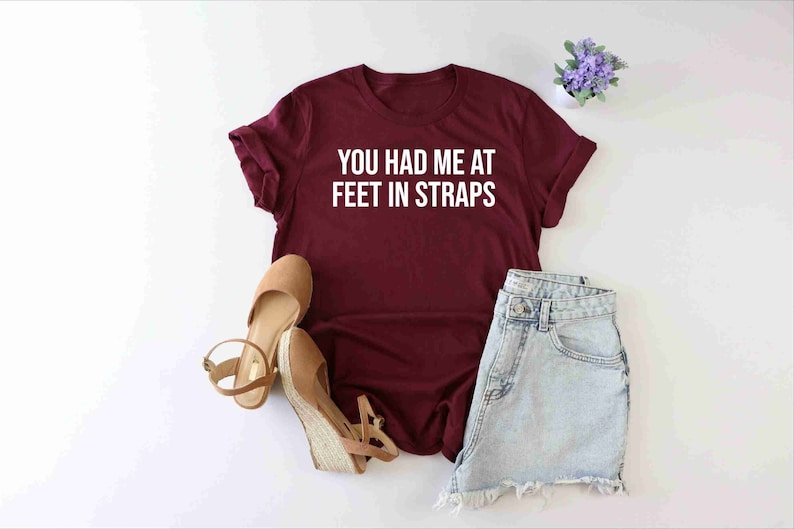 May include: Burgundy t-shirt with white text that reads "You had me at feet in straps". The shirt is laid flat on a white surface with a pair of brown sandals and denim shorts.