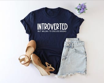 Könnte beinhalten: Ein marineblaues T-Shirt mit weißem Text, der "Introvertiert, aber bereit, über Bücher zu diskutieren" lautet.