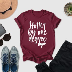 a casual outfit consisting of a burgundy t-shirt with the text "Hotter by one degree", a pair of black pants, a pair of sunglasses, a hat, and a small potted plant.