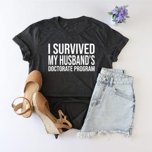 May include: A dark gray t-shirt with the text "I survived my husband's doctorate program" printed in white.