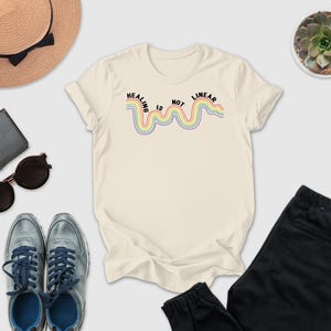 a casual outfit consisting of a white t-shirt with a rainbow-colored line, a pair of black pants, a pair of blue sneakers, sunglasses, a hat, and a small potted plant.