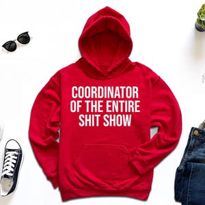 a red hoodie with the text "COORDINATOR OF THE ENTIRE SHIT SHOW" printed on it, along with a pair of jeans, sneakers, sunglasses, a watch, and a potted plant.