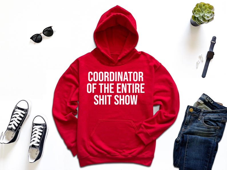 a red hoodie with the text "COORDINATOR OF THE ENTIRE SHIT SHOW" printed on it, along with a pair of jeans, sneakers, sunglasses, a watch, and a potted plant.