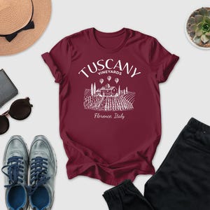 a collection of items, including a burgundy T-shirt with a graphic of a vineyard, sunglasses, a hat, a wallet, a pair of shoes, and a potted succulent.