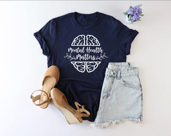 May include: A navy blue t-shirt with a white outline of a brain and the text "Mental Health Matters". The shirt is paired with light blue denim shorts and brown wedge sandals.