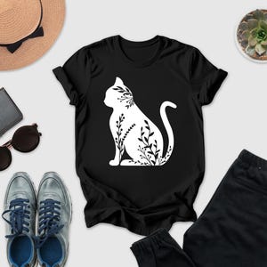 a casual outfit consisting of a black t-shirt with a white cat silhouette, a pair of gray sneakers, a pair of black pants, sunglasses, a hat, and a potted succulent plant.