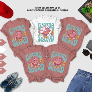 a collection of clothing items, including a white t-shirt with a pink and blue design, a pair of pink t-shirts with a similar design, and a pair of jeans. There are also a red hat, sunglasses, and a potted plant in the background.