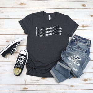 Puede incluir: Una camiseta gris oscuro jaspeada con el texto "Necesito m&aacute;s caf&eacute;" repetido tres veces en blanco.
