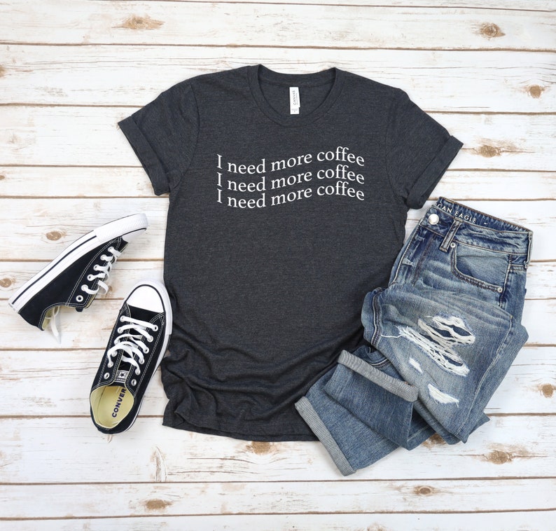 Puede incluir: Una camiseta gris oscuro jaspeada con el texto "Necesito m&aacute;s caf&eacute;" repetido tres veces en blanco.