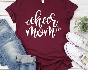 May include: A maroon t-shirt with the text "cheer mom" in white lettering with two small white hearts on either side of the text.