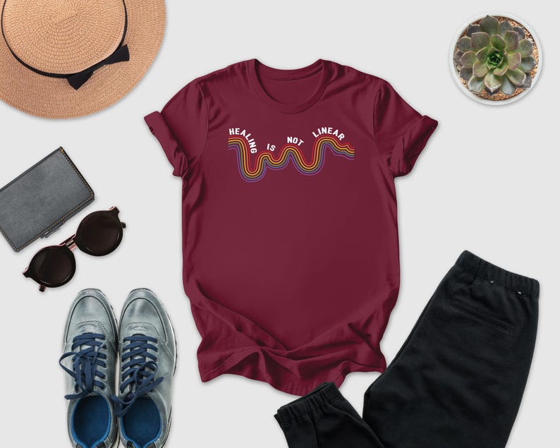 a casual outfit consisting of a burgundy t-shirt with a rainbow graphic, a pair of sunglasses, a hat, a wallet, a pair of shoes, and a potted plant.