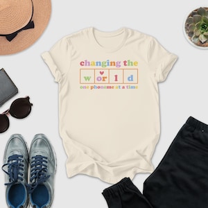 a casual outfit consisting of a white t-shirt with a colorful graphic, sunglasses, a hat, a wallet, a potted plant, and a pair of shoes.