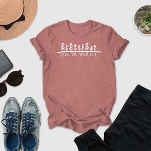 A pink t-shirt with the text "LIVE THE WILD LIFE" printed on it, along with a pair of sunglasses, a hat, a wallet, a potted succulent, and a pair of sneakers.