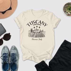 a casual outfit laid out on a white background, including a beige t-shirt with a graphic design, sunglasses, a hat, a wallet, a potted succulent, and a pair of black pants.