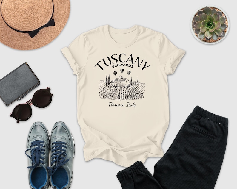 a casual outfit laid out on a white background, including a beige t-shirt with a graphic design, sunglasses, a hat, a wallet, a potted succulent, and a pair of black pants.