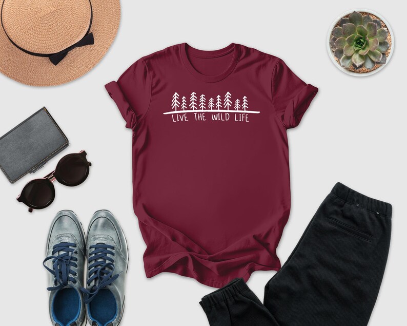 A maroon t-shirt with the text "LIVE THE WILD LIFE" printed on it, along with a pair of sunglasses, a hat, a wallet, a potted succulent, and a pair of sneakers.