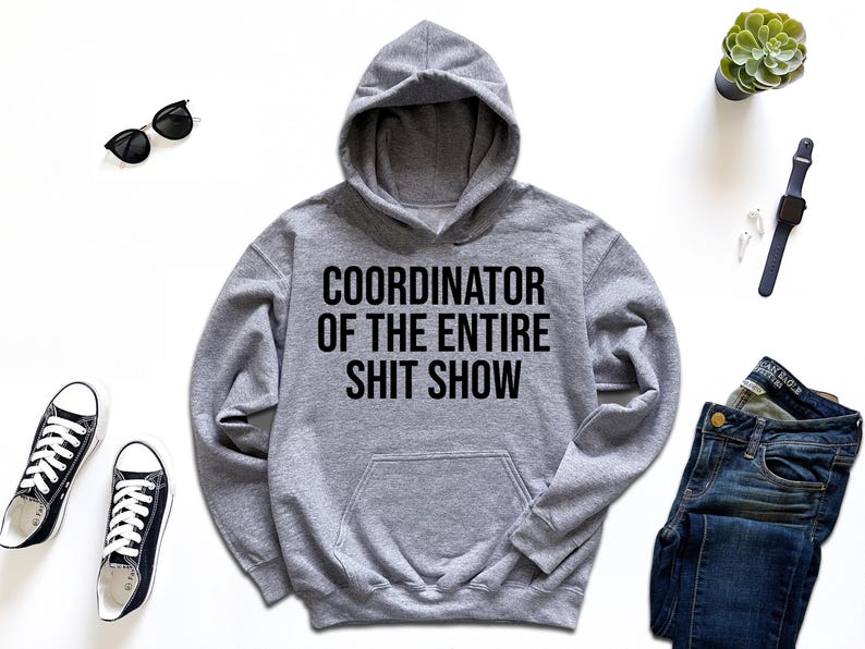 A gray hoodie with the text "COORDINATOR OF THE ENTIRE SHIT SHOW" printed on it, along with a pair of jeans, sneakers, a watch, sunglasses, and a potted plant.