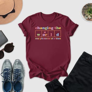 a variety of casual clothing and accessories, including a burgundy t-shirt with a colorful graphic, sunglasses, a hat, a wallet, a potted succulent, and a pair of sneakers.