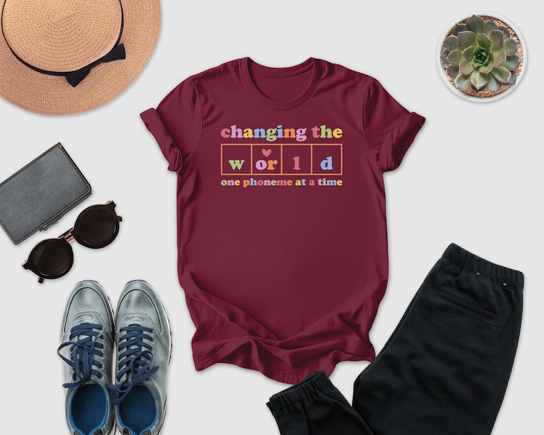 a variety of casual clothing and accessories, including a burgundy t-shirt with a colorful graphic, sunglasses, a hat, a wallet, a potted succulent, and a pair of sneakers.