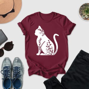 a casual outfit consisting of a burgundy t-shirt with a white cat silhouette, a pair of black pants, a pair of sunglasses, a hat, and a small potted succulent plant.