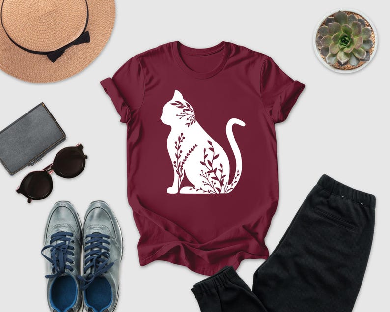 a casual outfit consisting of a burgundy t-shirt with a white cat silhouette, a pair of black pants, a pair of sunglasses, a hat, and a small potted succulent plant.