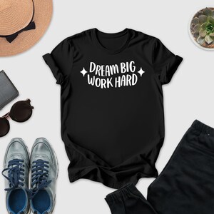 a casual outfit consisting of a black t-shirt with the text "DREAM BIG WORK HARD" printed on it, a pair of gray sneakers, a pair of sunglasses, a hat, and a potted succulent plant.