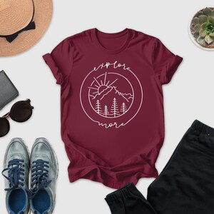 a casual outfit consisting of a burgundy t-shirt with a graphic design, a pair of sunglasses, a hat, a wallet, a pair of shoes, and a potted succulent plant.