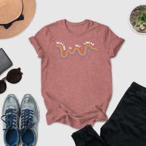 a casual outfit consisting of a pink t-shirt with a rainbow-colored line, a pair of black pants, a pair of blue sneakers, sunglasses, a hat, and a small potted plant.