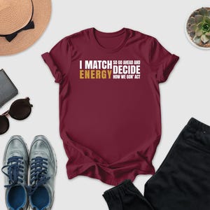 a casual outfit consisting of a burgundy t-shirt with a humorous text, a pair of gray sneakers, a hat, sunglasses, a wallet, a potted plant, and a pair of black pants.