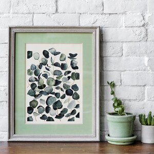 May include: A framed watercolor painting of eucalyptus leaves in shades of green and blue on a white background. The frame is a light gray with a green mat.
