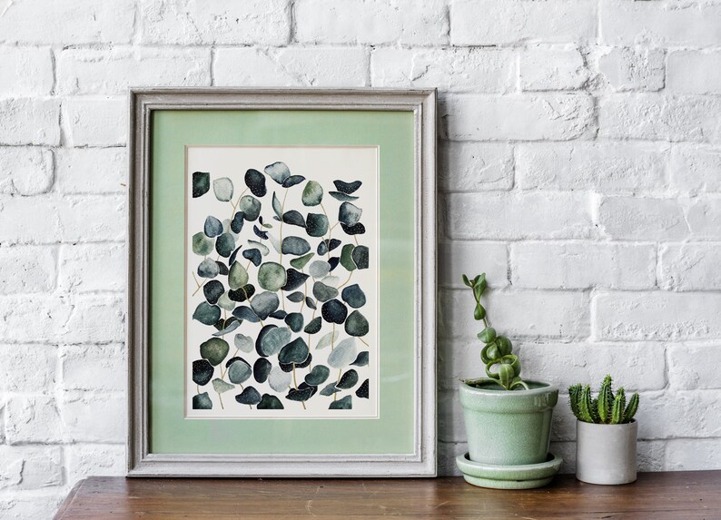 May include: A framed watercolor painting of eucalyptus leaves in shades of green and blue on a white background. The frame is a light gray with a green mat.