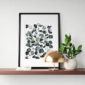 May include: A framed watercolor print of eucalyptus leaves in shades of green and blue, a gold dome lamp, and a potted green plant on a brown shelf.